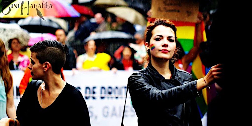 A young woman with red hair pulled back and wearing a black jacket holds a sign in both hands. She has a rainbow flag painted on her left cheek. The background behind her is out of focus, but filled with rainbow umbrellas clustered together. 