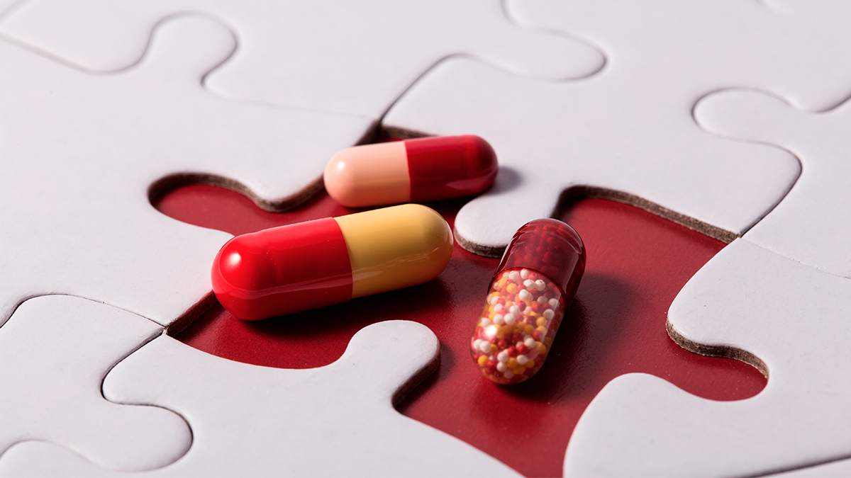 A close up of white puzzle pieces fitting together with one piece missing showing a red background. Three capsules of medication from the Life sciences sector fill the empty spot.