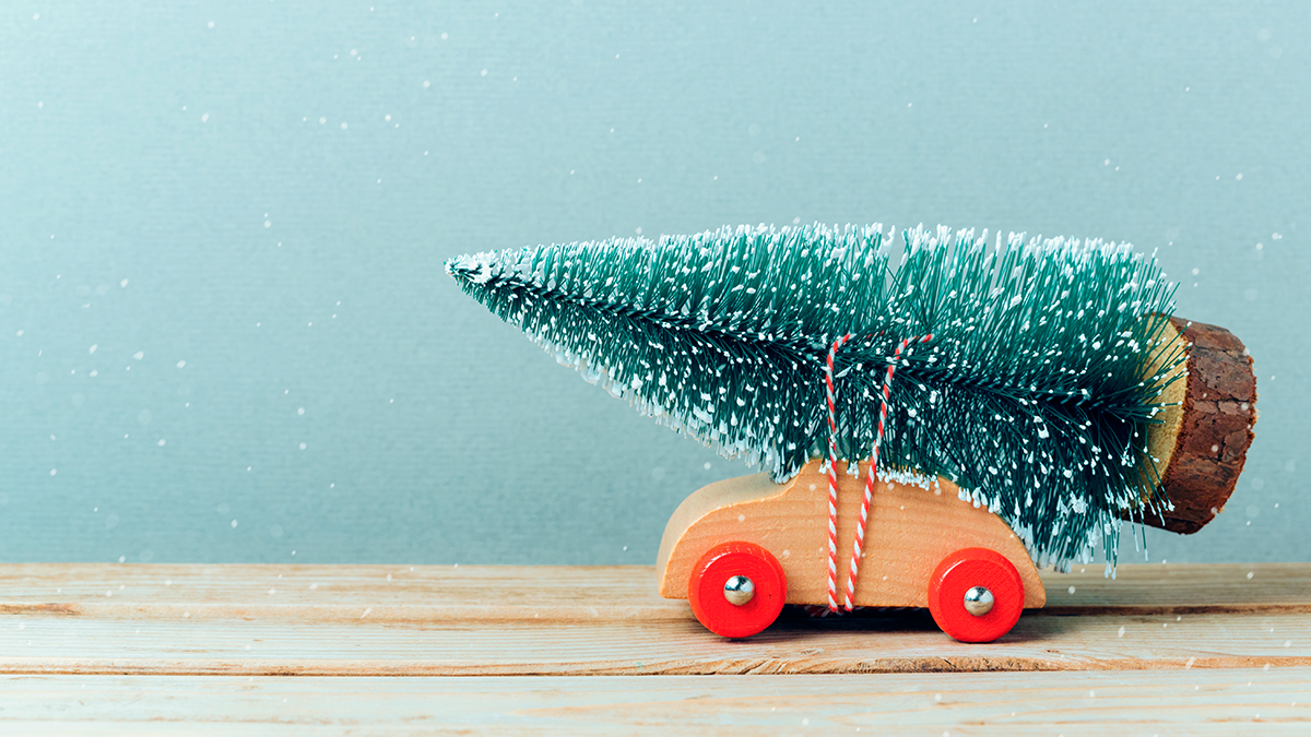Little toy wooden car with toy Christmas tree tied to its roof appears to be traveling from right side of image to left on a wooden table top.